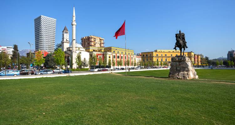 Stadtplatz mit einer Statue, Flagge und Gebäuden einschließlich eines Uhrenturms.