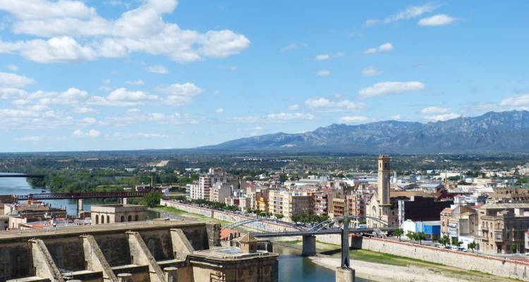 Ein Blick auf eine Stadt entlang eines Flusses mit Bergen im Hintergrund unter einem blauen Himmel.
