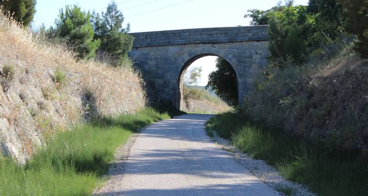 Eine kleine Steinbrücke über einen Weg, umgeben von grasbewachsenen Böschungen an einem sonnigen Tag.