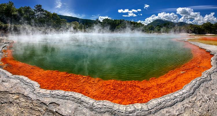 Farbenfrohe geothermale Heißquelle mit aufsteigendem Dampf unter blauem Himmel.