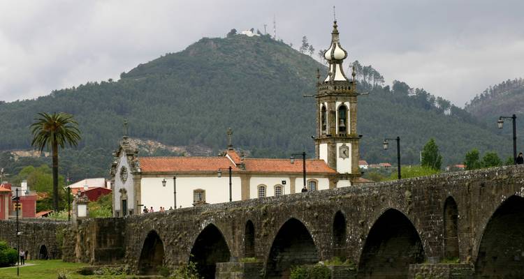 Medieval stone bridge with multiple arches spanning a calm river with a church tower and forested mountain backdrop.
