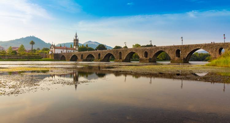 Sunlit stone bridge and church reflected in calm river waters with a blue-gold sky.