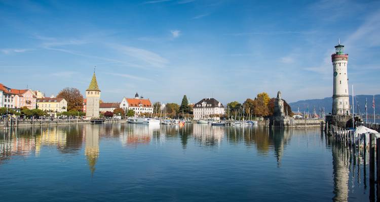 Façade panoramique au bord de l'eau avec phare, tours historiques et bateaux amarrés se reflétant dans un lac calme.