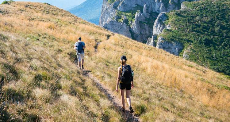 Two hikers with backpacks ascend a grassy alpine trail toward rugged cliffs.