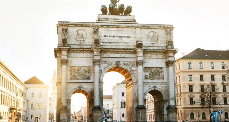 Der sonnige Siegestor-Triumphbogen erhebt sich über einen breiten Boulevard, der von historischen Münchner Gebäuden flankiert wird.