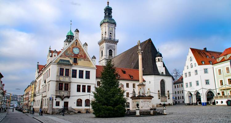 Ruhiger bayerischer Marktplatz mit historischen Kirchen, Pastellgebäuden und einem zentralen Denkmal unter bewölktem Himmel.