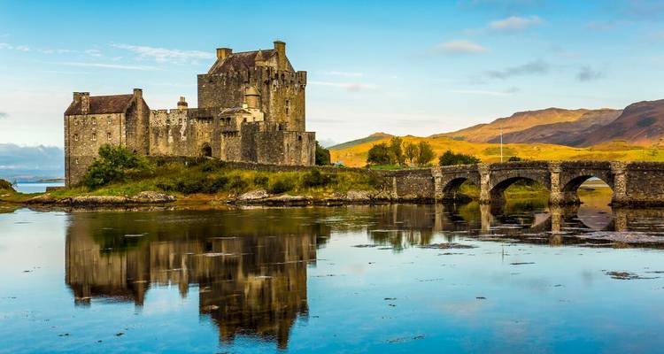 Le château d'Eilean Donan se reflétant dans les eaux calmes avec un pont en pierre et les Highlands au loin.