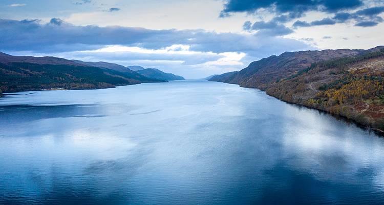 Vista aérea de un largo loch azul flanqueado por las Highlands boscosas bajo un cielo nublado.