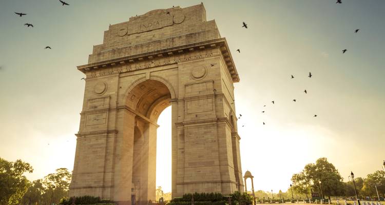 Monumento India Gate a contraluz del sol matutino con pájaros volando en cielo dorado y brumoso.