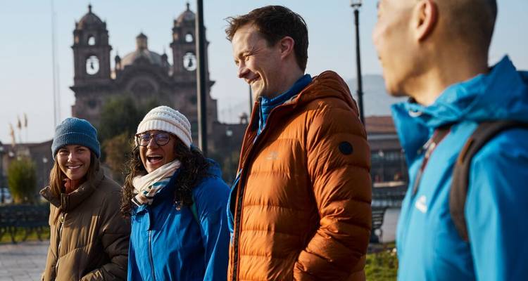 Grupo de viajeros en chaquetas abrigadas pasean y conversan en la Plaza de Armas de Cusco con una catedral detrás.