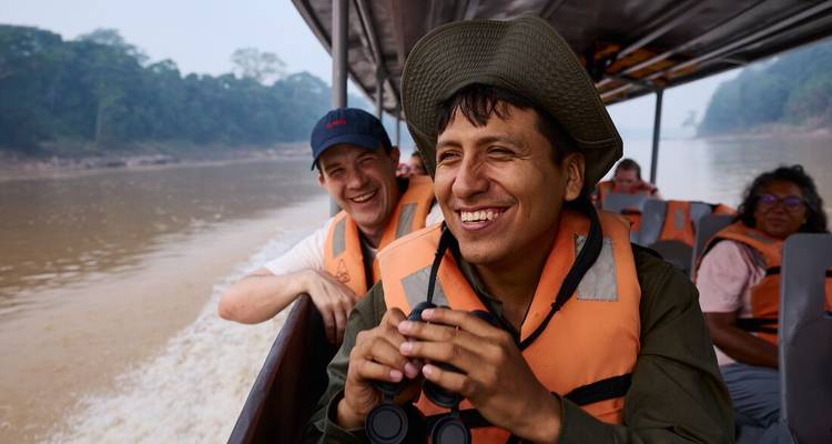 Viajeros sonrientes que llevan chalecos salvavidas viajan en una lancha motorizada por un río amazónico ancho y fangoso.