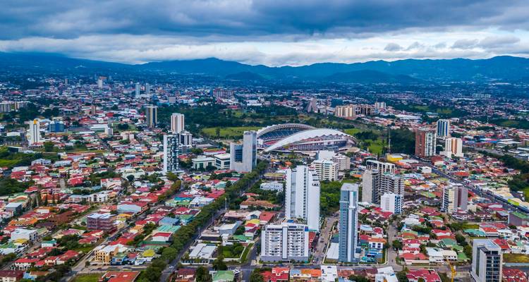 Panorama aérien de la ville de San José montrant un stade moderne, des gratte-ciel et les montagnes environnantes sous un ciel nuageux.