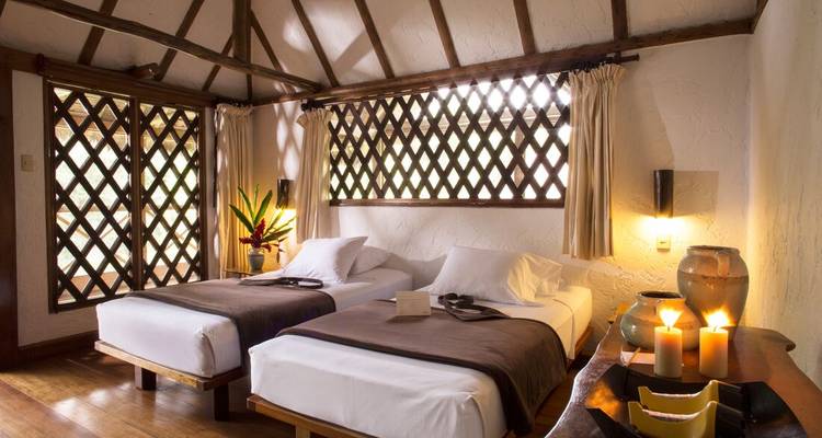 Rustic twin-bed jungle lodge room with wooden beams, candlelight and patterned lattice windows.