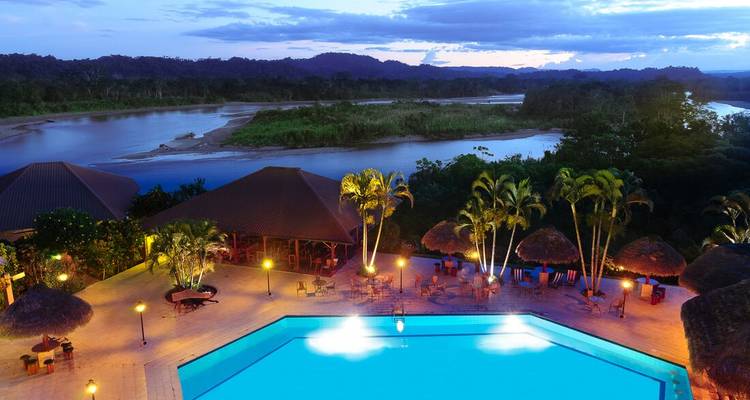 Aerial evening view of a riverside jungle lodge with illuminated pool and palm trees beside calm water.