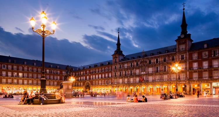 Ambiente nocturno en la Plaza Mayor de Madrid, con gente relajándose alrededor de la farola central y los edificios rojos con arcadas iluminados cálidamente contra un cielo crepuscular.