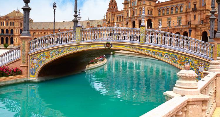 Puente decorativo de azulejos que atraviesa el canal turquesa de la Plaza de España en Sevilla con arquitectura ornamentada al fondo.
