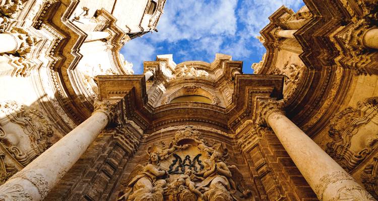 Vista dramática hacia arriba de la fachada barroca de la Catedral de Valencia con columnas imponentes y tallados intrincados contra un cielo azul profundo.