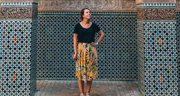 Mujer de pie frente a una pared de azulejos coloridos, posiblemente marroquí.
