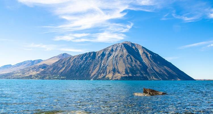 Un lago de un azul cristalino con una orilla rocosa y una montaña empinada detrás bajo nubes ligeras.