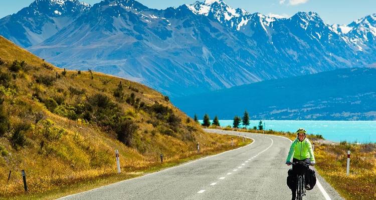Un ciclista con ropa brillante anda en bicicleta por una carretera sinuosa junto a un lago turquesa con los Alpes del Sur nevados de fondo.