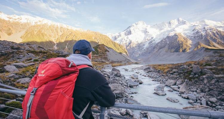 Excursionista con mochila roja admira un río glacial que serpentea a través de un valle montañoso nevado en el Parque Nacional Aoraki.