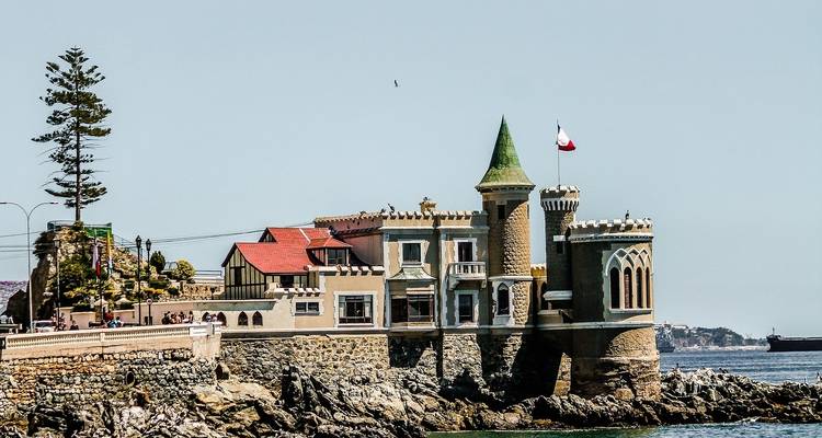 A castle-like building on a rocky oceanfront with a distant city skyline.