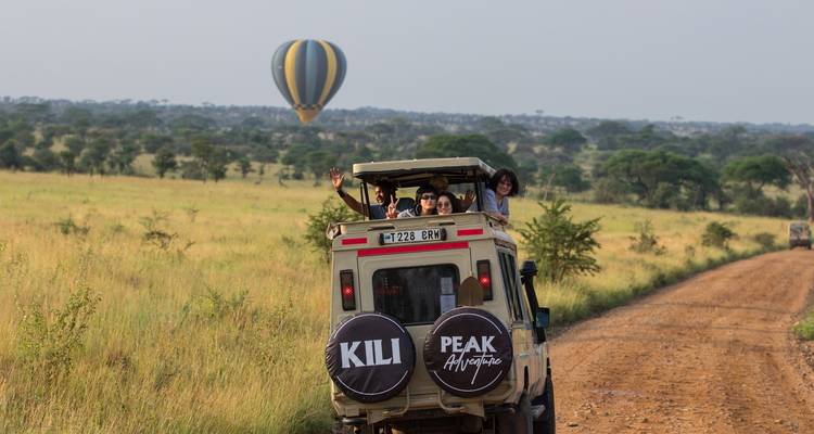 Véhicule de safari avec des touristes qui saluent de la main, et une montgolfière au loin.