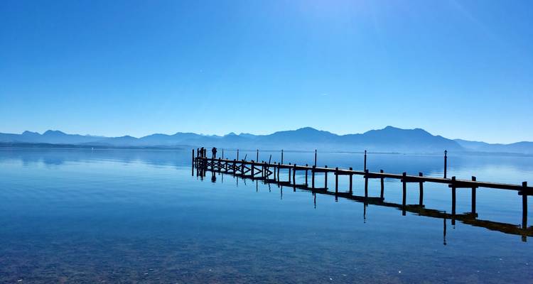 Langer Holzsteg, der sich über den spiegelglatten See erstreckt, mit zwei Personen, die am fernen Ende stehen, Bergsilhouetten im Hintergrund unter einem klaren blauen Himmel.
