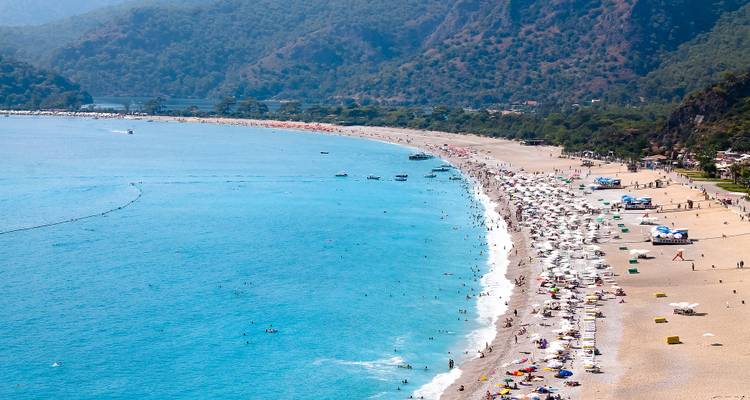 Plage avec une eau bleue claire et des collines environnantes, bondée de gens et de parasols.