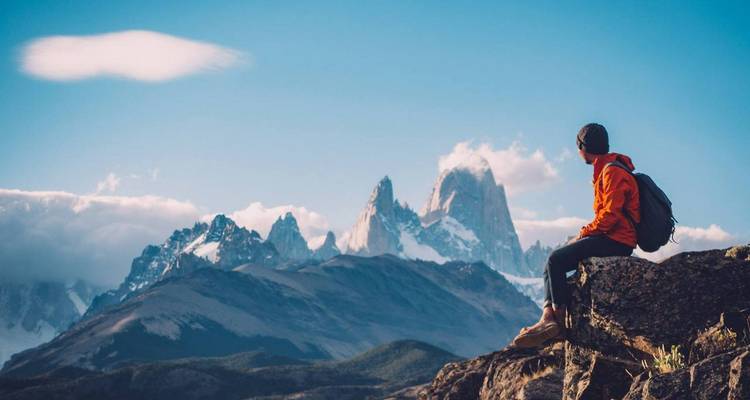 Jonge wandelaar in oranje jas zittend op een rots, bewonderend de scherpe besneeuwde toppen van Mount Fitz Roy.