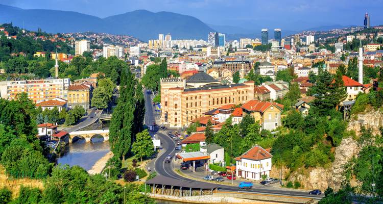 Panoramablick auf Sarajevo mit Fluss, Gebäuden und umliegenden Hügeln.