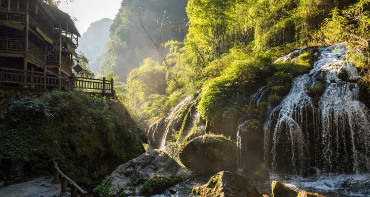 Malerische Aussicht auf einen Wasserfall und üppiges Grün mit einem traditionellen Gebäude an der Seite.