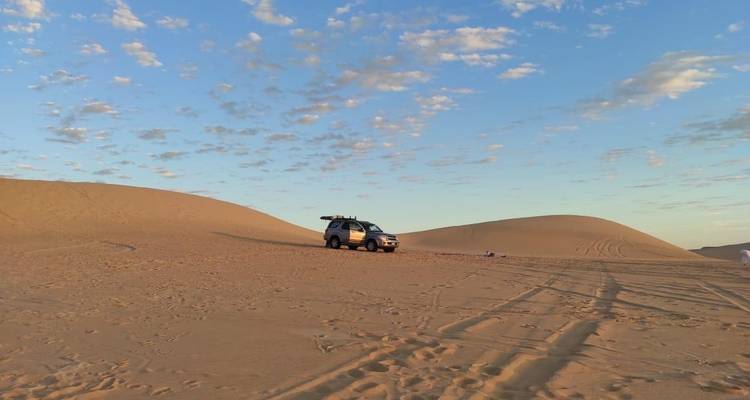 SUV geparkeerd op een woestijnduin onder een blauwe lucht met verspreide wolken.