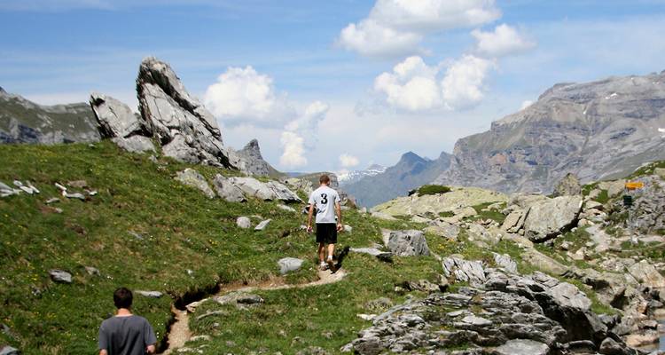 Excursionistas siguiendo un estrecho sendero de montaña entre un paisaje alpino rocoso bajo un cielo parcialmente nublado