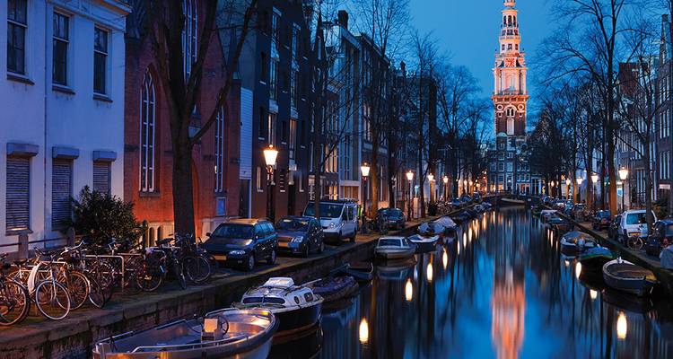Vista al atardecer a lo largo de un canal de Ámsterdam con bicicletas, botes y la torre de una iglesia suavemente iluminada reflejada en el agua.