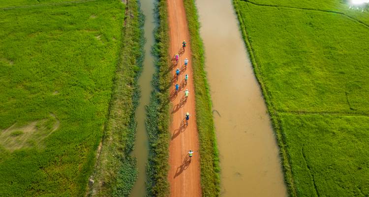 Vue aérienne de cyclistes sur un sentier de terre entre des champs verts luxuriants et l'eau.