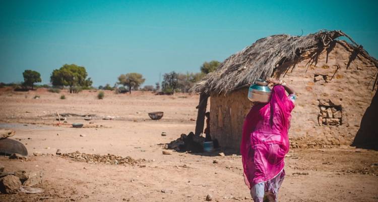 Femme en tenue traditionnelle rose portant un pot sur sa tête près d'une maison rustique.