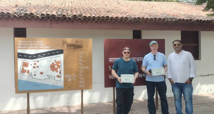 Three visitors holding certificates beside an outdoor Gandhi Ashram site map board