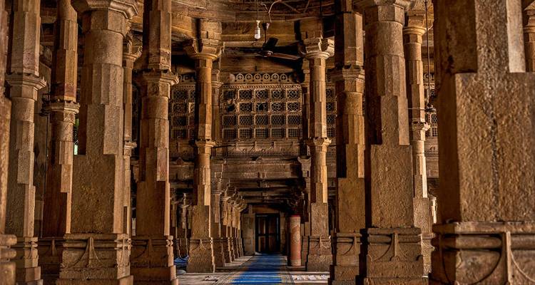 Long hall of towering stone pillars with patterned carpet leading to a doorway in an Indian mosque