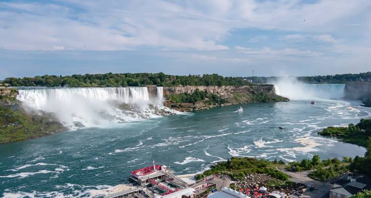 Wide aerial of Niagara Falls showing both American and Horseshoe Falls with tour boats navigating the rapids.