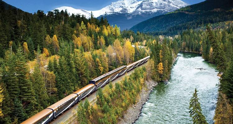 Luxury sightseeing train snaking along a forested river valley with colourful autumn trees and distant mountains.