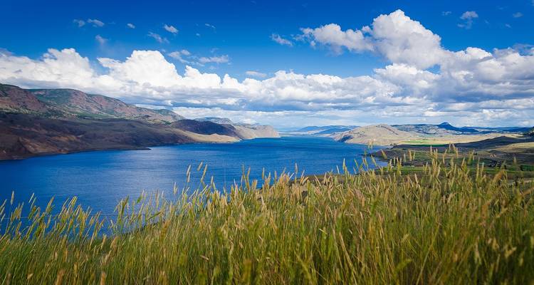 Blue lake stretching through rolling semi-arid hills and tall grasses beneath a dramatic cloud-filled sky.
