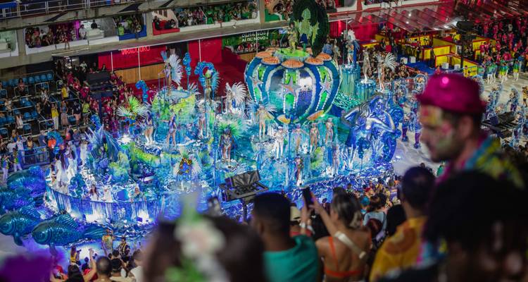 Carro alegórico con tema azul iluminado con performers pasando frente a los espectadores en el Sambódromo por la noche