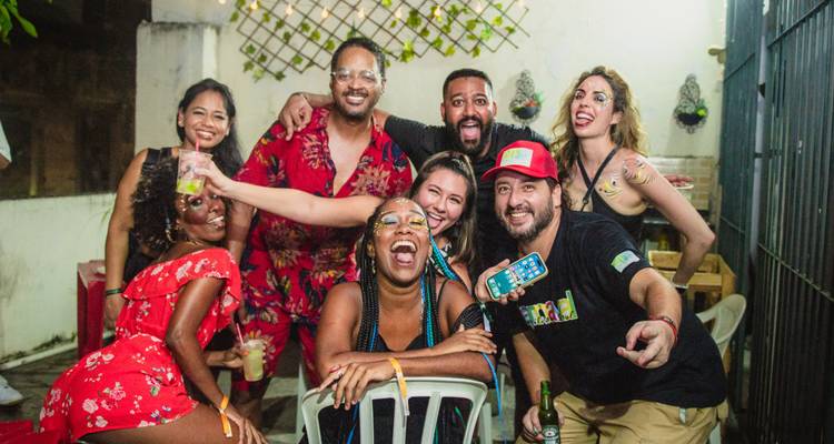 Un grupo animado de jóvenes adultos riendo y posando juntos en una fiesta al aire libre, sosteniendo bebidas y vistiendo atuendos coloridos.