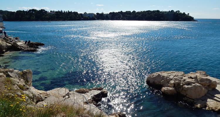 La luz del sol brilla en el Mar Adriático cerca de la costa rocosa y la península distante cubierta de árboles.