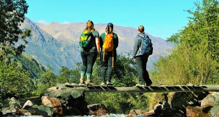 Drie wandelaars op een houten brug met uitzicht op bergen.