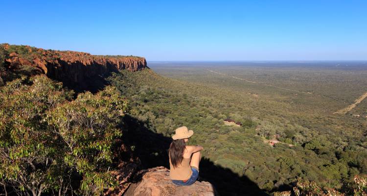 Een persoon zit op een klif met uitzicht op uitgestrekte wildernis.