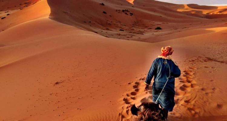 Een persoon in traditionele kleding die een kameel door zandduinen leidt.