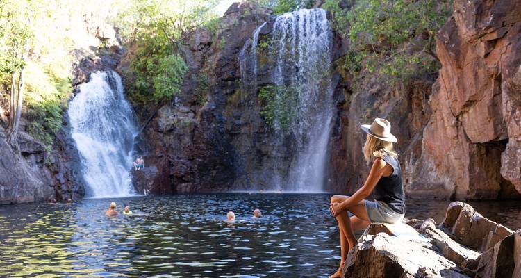 Des chutes d'eau jumelles se jettent dans un bassin limpide où des nageurs se détendent et un voyageur en chapeau observe depuis les rochers