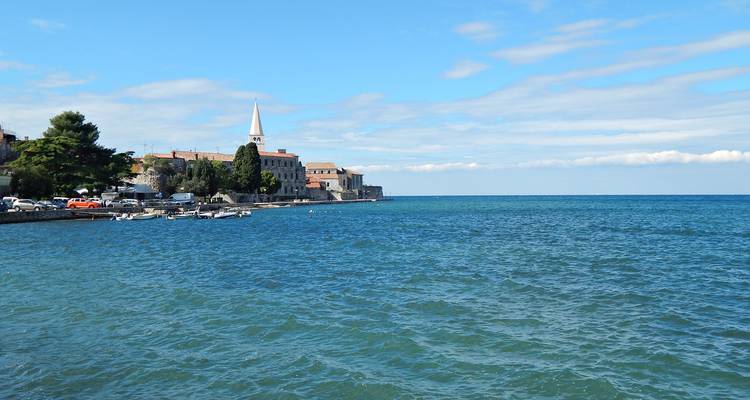 Les eaux bleues de l'Adriatique avec une ville côtière, un clocher d'église et une petite marina sous des nuages épars.